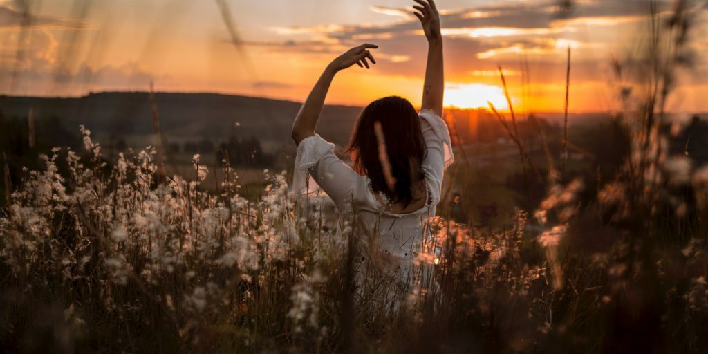 woman-wearing-white-dress-raising-her-two-hands-surrounded-white-petaled-flowers-during-sunset
